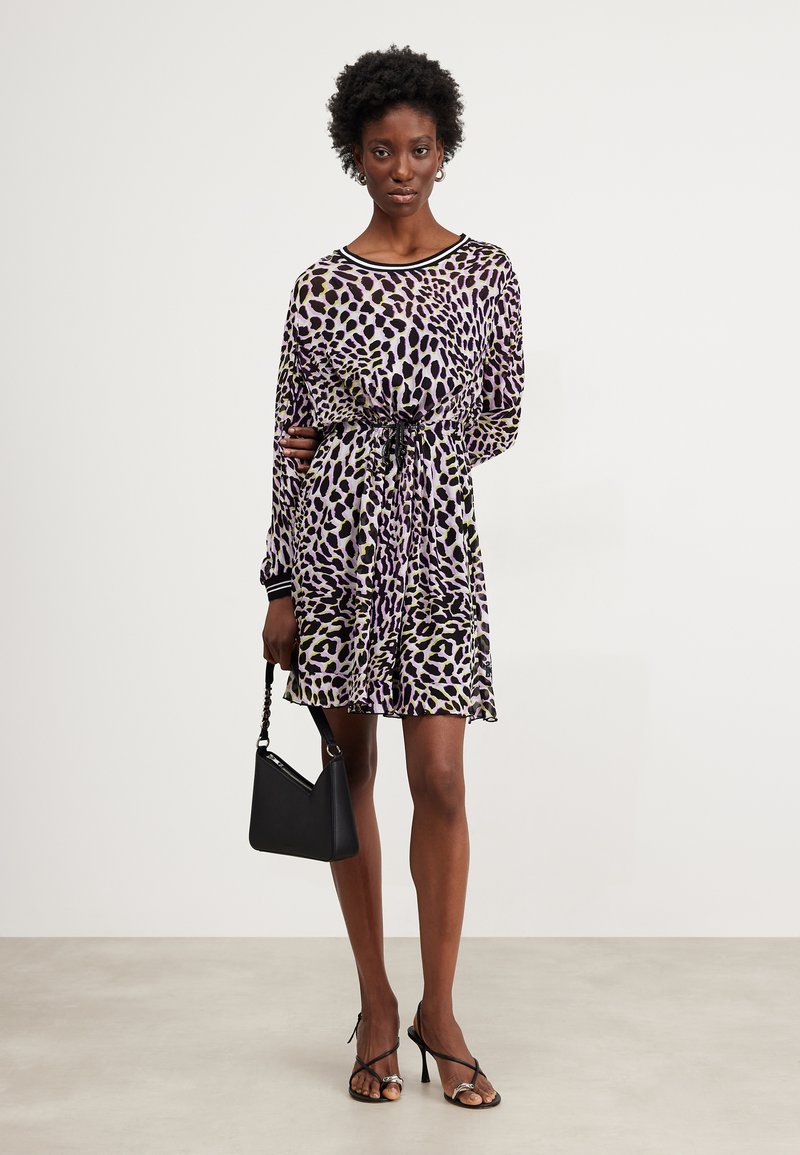 Long-sleeve dress in purple with a leopard print pattern, cinched at the waist with a black tie. Paired with a black handbag and strappy heels.