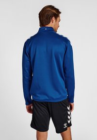 Giacca sportiva blu con colletto alto e righe bianche sulle spalle, abbinata a pantaloni corti neri con dettagli a chevron bianchi. Tessuto liscio.