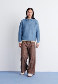 Μπλε πουλόβερ με υφή, κολάρο και τσέπη, συνδυασμένο με καφέ καρό παντελόνι φαρδιάς γραμμής και κρέμα παπούτσια, σε απλή φόντο.