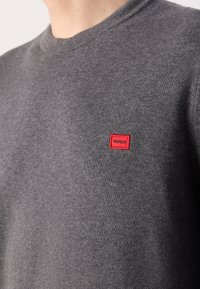 Gros plan sur une personne portant un pull en maille gris avec une petite étiquette rectangulaire rouge portant l'inscription "HUGO" sur la poitrine.