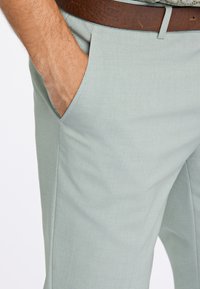 Vīrieša roka gaiši pelēku biksu kabatā, redzams brūns ādas jostas siksna un rakstains krekls virs jostasvietas.