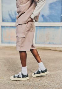 Personne portant un short cargo beige et une chemise boutonnée assortie, des baskets noires et blanches, avec une main dans la poche, debout sur le trottoir.