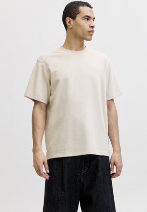 Man draagt een effen beige T-shirt met korte mouwen en losse zwarte broek, kijkt naar rechts tegen een lichtgrijze achtergrond.