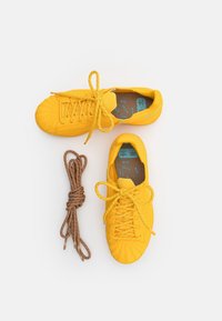 Ljusa gula sneakers med ovandel i texturerat tyg, rund tå och bruna skosnören bredvid. Insidan har en blå innersula.