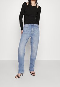 Haut noir près du corps à manches longues avec détail épaule découpée, associé à un jean bleu clair taille haute à jambes droites et des talons noirs à brides.