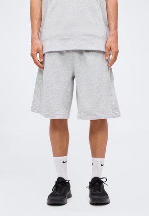 Mężczyzna ubrany w jasnoszorty do kolan, białe skarpety Nike i czarne buty sportowe, stojący na jednolitym białym tle.