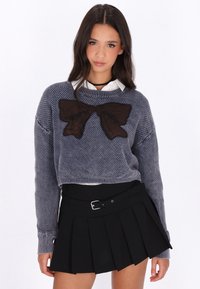 Pulover tricotat albastru, scurt, cu un design mare de fundă maro în față, asociat cu o fustă neagră plisată dotată cu un bretea.