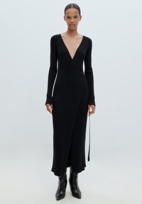 Schwarzes, geripptes Strickkleid mit langen Ärmeln und tiefem V-Ausschnitt, mit einem Wickeldesign und knöchellangem Saum. Kombiniert mit spitzen schwarzen Stiefeln.