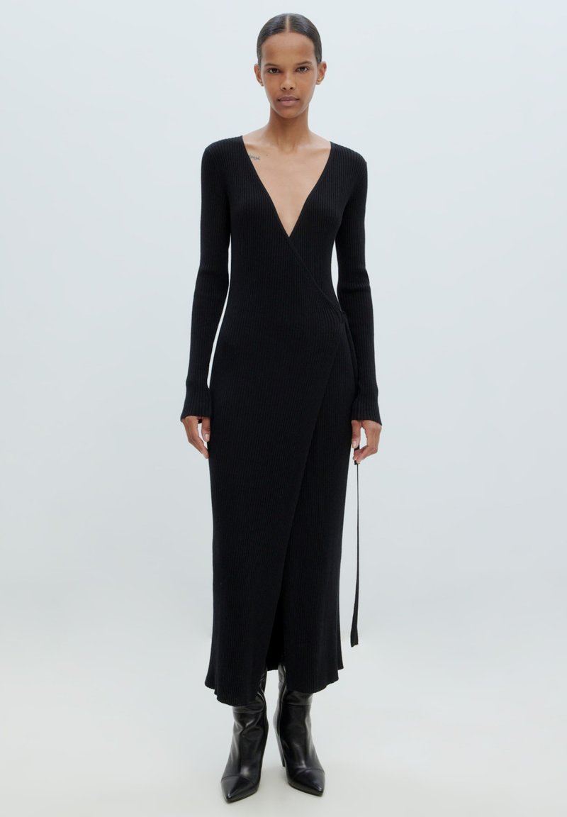 Schwarzes, geripptes Strickkleid mit langen Ärmeln und tiefem V-Ausschnitt, mit einem Wickeldesign und knöchellangem Saum. Kombiniert mit spitzen schwarzen Stiefeln.
