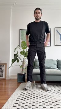 Black cargo pants with side pockets, a black t-shirt, and gray sneakers. The setting includes a green plant and a light-colored couch.