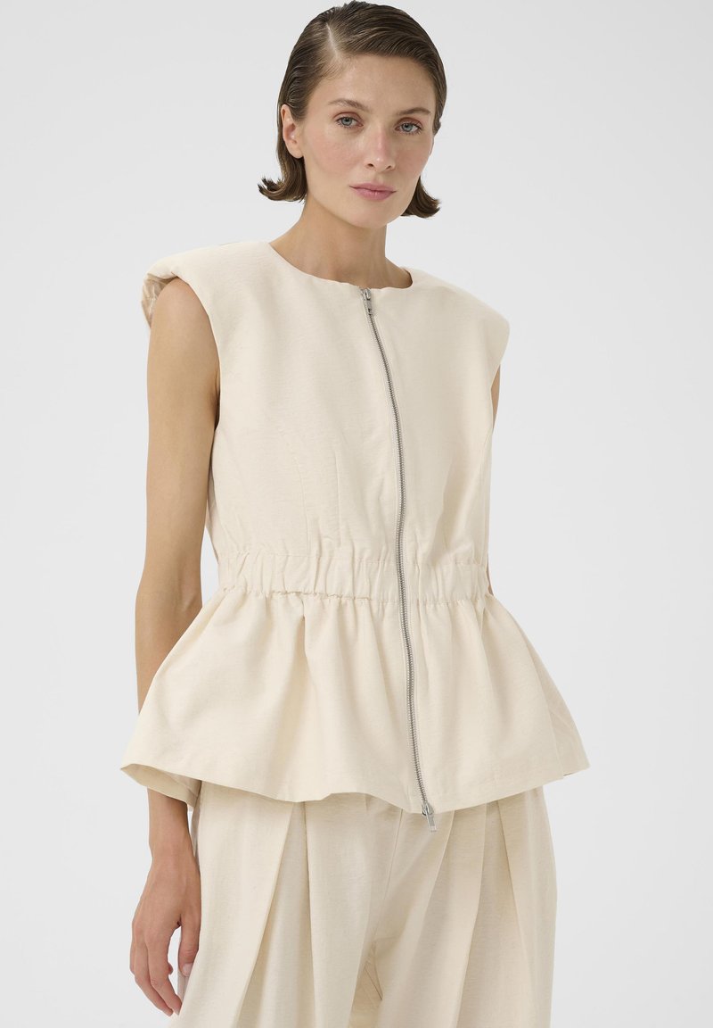 Mujer de cabello corto vistiendo un top sin mangas beige con cremallera y peplum, junto con pantalones anchos a juego, sobre un fondo liso.