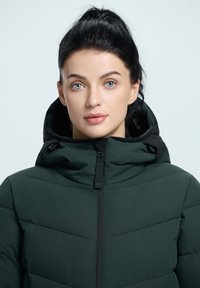 Chaqueta acolchada verde oscuro con capucha, con cierre de cremallera y sutiles patrones de costura en diagonal, hecha de un material liso y texturizado.