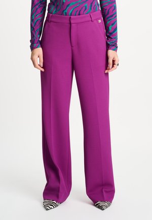 Vrouw draagt hooggetailleerde, wijd uitlopende paarse broek, gecombineerd met zwart-witte zebraprint spitse schoenen en een langarmige top met patroon.