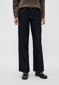 Τζιν παντελόνια σε στυλ wide-leg από σκουρόχρωμο denim με αντίθετες ραφές, διαθέτοντας μέση μεσαίου ύψους και κλασικό σχεδιασμό με πέντε τσέπες. Φορεμένα με καφέ πουλόβερ και μπότες.