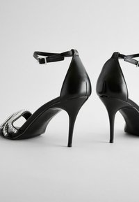 Sandale negre cu toc înalt, cu un finisaj din piele lucioasă, având o curea subțire la gleznă și un accent decorativ cu pietre strălucitoare în față.