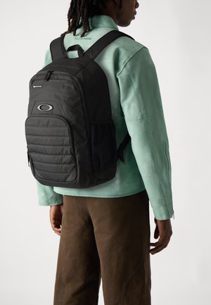 Sac à dos noir avec texture matelassée et grande poche avant, doté d'une poche latérale en mesh et d'un logo. Porté sur une veste verte.