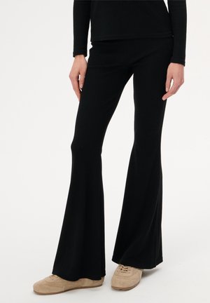 Osoba ubrana w czarne rozszerzane spodnie i beżowe buty na sznurowadła, stojąca na tle jednolitej białej ściany.