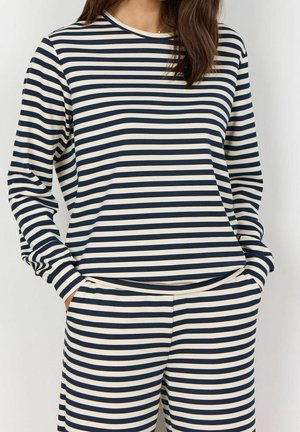 Woman wearing a long-sleeve top and pants with horizontal navy and white stripes, hands in pockets, against a plain light background.