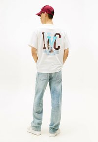 Witte katoenen t-shirt met NYC-afbeelding in blauw, rood en wit. Lichtblauwe denimjeans, witte sneakers en een bordeauxrode pet.