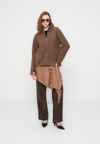 Καφέ φλις μπουφάν με φερμουάρ και ψηλό γιακά, στρωτό μπεζ τοπ με λεπτομέρειες βολάν, καφέ παντελόνι με φαρδιά πόδια και μαύρα παπούτσια με τακούνι.