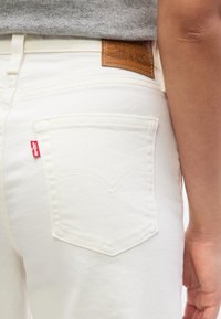 Άτομο που φοράει λευκά τζιν Levi's με την κόκκινη ετικέτα της μάρκας στην πίσω τσέπη και καφέ δερμάτινο ταμπελάκι στη ζώνη μέσης.