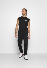 Canotta nera senza maniche con logo bianco, abbinata a pantaloni sportivi neri con una striscia grigia e scarpe da ginnastica bianche con accenti colorati.