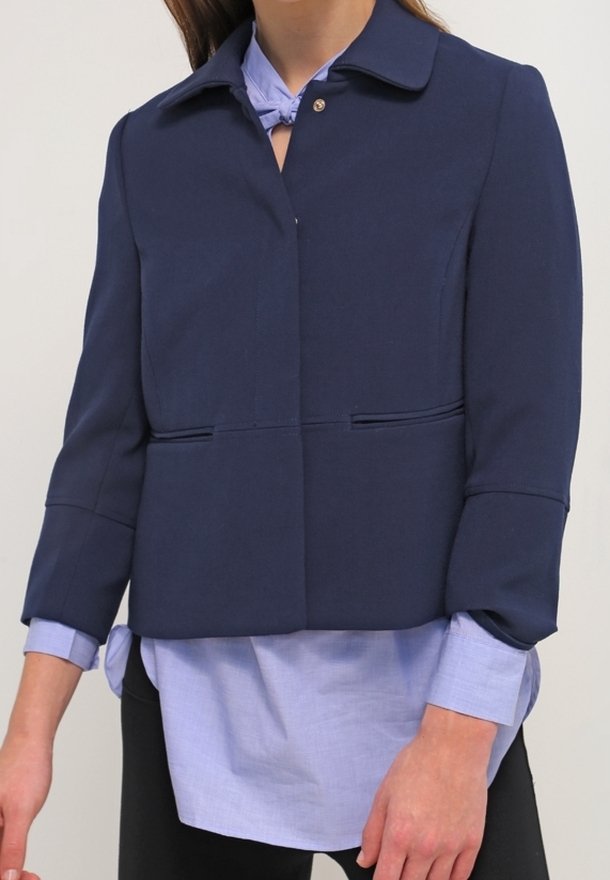 Femme portant une veste courte bleu marine par-dessus une chemise à boutons bleu clair et un pantalon noir, debout devant un fond uni.