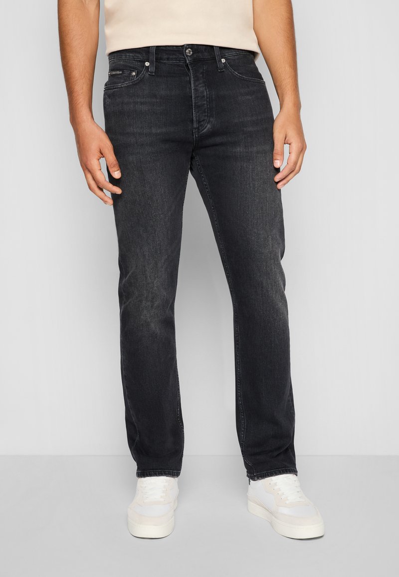 Jean en denim noir avec une coupe droite, caractérisé par un style à cinq poches et une texture légèrement décolorée, porté avec des baskets blanches.