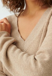 Pull beige en maille avec un col en V et une texture côtelée. Manches longues avec une coupe décontractée, accents subtils de scintillement au niveau du col.