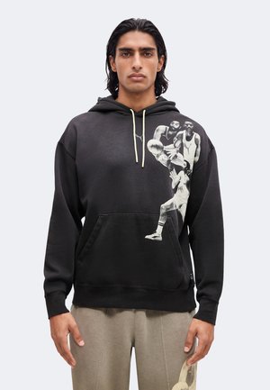 Homme aux cheveux foncés portant un sweat à capuche noir avec une impression en noir et blanc d’un joueur de basket-ball sur la poitrine et un pantalon beige avec un imprimé sur la cuisse.