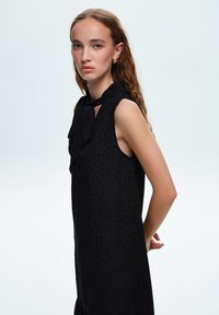 Svart klänning utan ärmar med en texturerad leopardmönster, med en rosett vid halsringningen och en avslappnad silhuett.