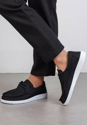 Personne portant des chaussures slip-on noires avec des semelles blanches et un pantalon noir, debout à l'intérieur sur un sol gris.