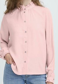 Vrouw in een lichtroze geknoopte blouse met gerimpelde kraag en manchetten, gecombineerd met lichtblauwe jeans, met een hand in haar zak.