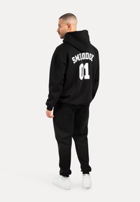 Czarny zestaw z bluzy i spodni wykonany z miękkiego materiału. Bluza ma na plecach napis "SMILODON 01" w białym kolorze. Dobrane do białych sneakersów.