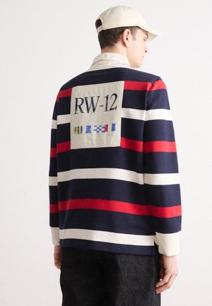 Chemise à manches longues rayée bleu marine, rouge et blanche avec une patch beige affichant "RW-12" et des symboles colorés au dos, fabriquée en coton.