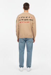 Felpa beige con testo grafico sul retro, colletto a girocollo e polsini a coste. Abbinata a jeans chiari e testurizzati, e sneaker bianche.