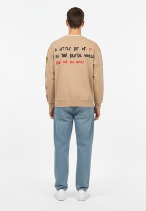 Beige sweatshirt met grafische tekst op de achterkant, ronde halslijn en geribde manchetten. Gepaard met lichtblauwe, gestructureerde jeans en witte sneakers.