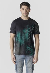 Mees, kes kannab musta ja rohelist graafilise mustriga T-särki ning helesiniseid cargo-tüüpi teksaseid, seisab helehalli ühevärvilise tausta ees.