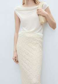 Haut blanc drapé avec un col large associé à une jupe crayon crème texturée présentant un motif en relief, accessoirisé d'un sac en osier.