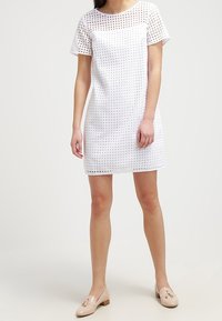 White short-sleeve dress with a grid pattern, made from lightweight material. Paired with beige flat shoes featuring a decorative accent.