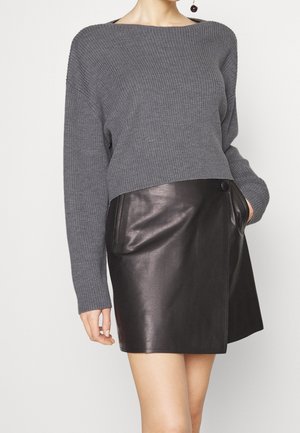 Frau trägt einen grau gerippten Pullover und einen schwarzen Mini-Lederrock mit Knopfdetail und Seitentaschen.
