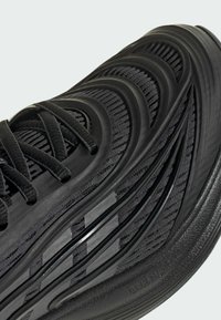 Zapatilla deportiva negra con parte superior texturizada y detalles en capas, forma redondeada y cordones planos. Suela de caucho duradera con detalle de marca.