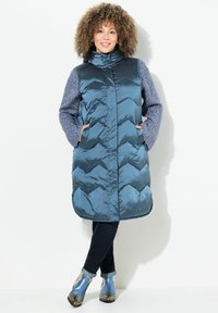 Padded teal farbiger langer Weste mit hohem Kragen, Zickzackmuster und blauen Strickärmeln. Kombiniert mit dunklen Jeans und metallic blauen Stiefeletten.