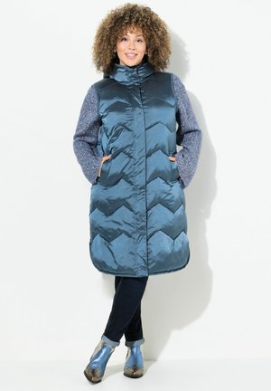 Padded teal farbiger langer Weste mit hohem Kragen, Zickzackmuster und blauen Strickärmeln. Kombiniert mit dunklen Jeans und metallic blauen Stiefeletten.