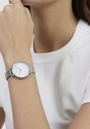 Montre ton argent avec un cadran rond blanc et un bracelet en acier inoxydable à maille. Présente des marqueurs d'heure minimalistes et une surface lisse et élégante.