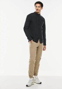 Schwarze Hemd mit Stehkragen, kombiniert mit beigen Chinos und weißen Sneakers, vor einem weißen Hintergrund stehend.