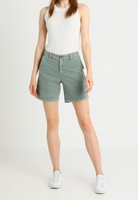 Shorts en coton vert clair à coupe décontractée, poches avant et fermeture à bouton, associés à un haut en tricot blanc sans manches et des baskets blanches.
