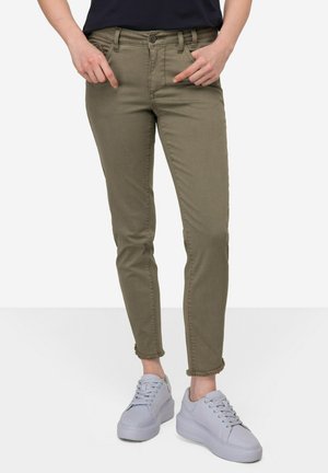 Olivgrüne Skinny-Jeans mit fransigem Saum, Fünf-Taschen-Design und passender Naht, kombiniert mit hellgrauen Plateausneakers.