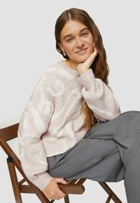 Heller rosa Pullover mit strukturierten weißen Mustern, Rundhalsausschnitt und gerippten Bündchen. Trägt dazu graue Hosen und sitzt auf einem Holstuhl.