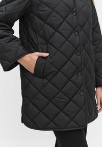 Svart quiltad kappa med ett diamantmönster, med en framlåda, runda knappar och långa ärmar. Tillverkad av ett glatt syntetiskt material.